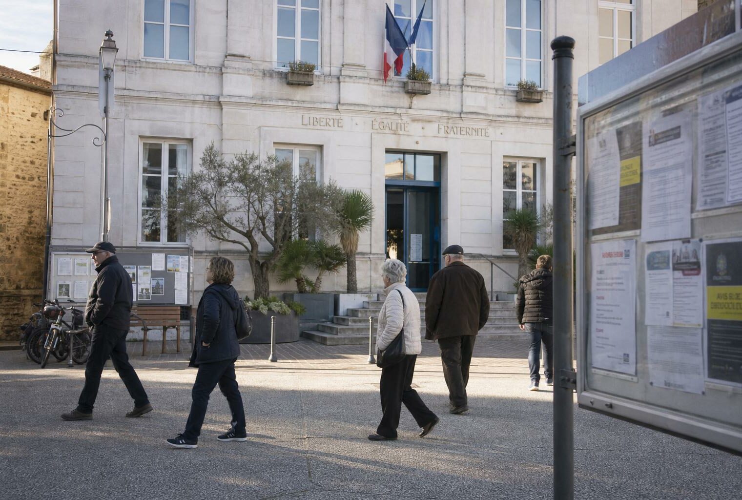 Place de mairie en ville française, passants anonymes et panneau d’affichage flou dans une lumière de journée claire.