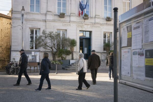 Place de mairie en ville française, passants anonymes et panneau d’affichage flou dans une lumière de journée claire.