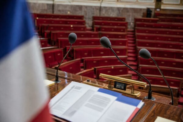 Salle de presse de l’Assemblée nationale avec pupitre vide, micros et sièges rouges, illustrant le débat sur l’aide à mourir.