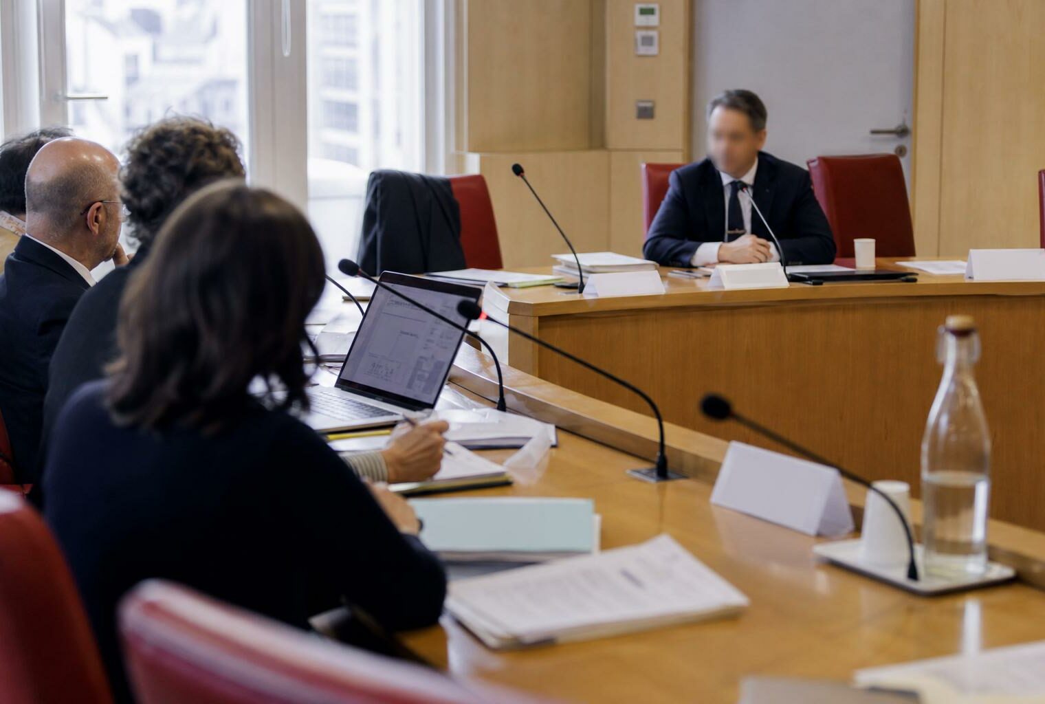 Audition en commission parlementaire avec micros, dossiers ouverts et silhouettes anonymes dans une salle institutionnelle lumineuse.