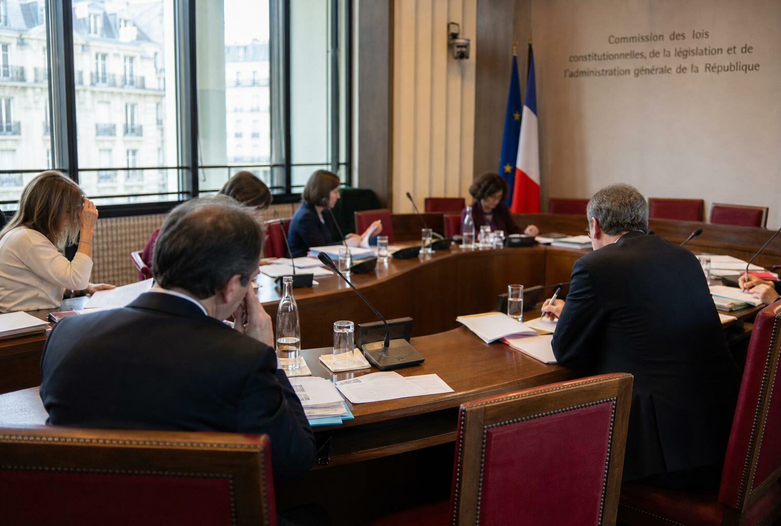 Audition parlementaire en commission avec micros, dossiers ouverts et silhouettes anonymes dans une salle lumineuse