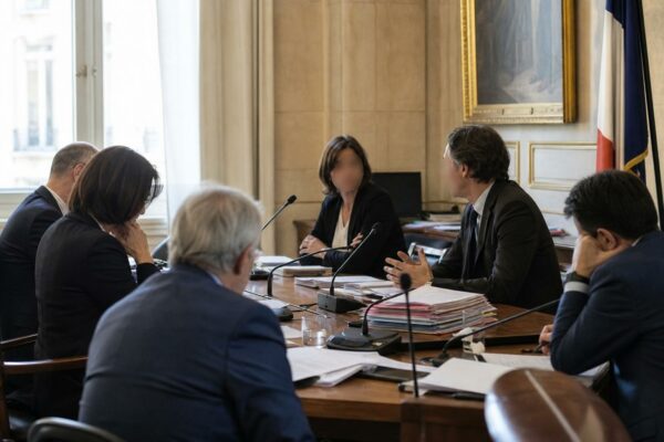 Audition parlementaire en France avec micros, dossiers ouverts et silhouettes anonymes dans une salle lumineuse