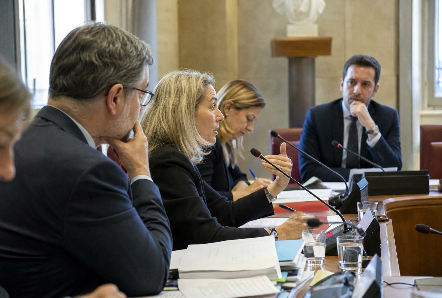 Audition parlementaire dans une salle institutionnelle avec micros, dossiers et silhouettes anonymes autour d’une table.