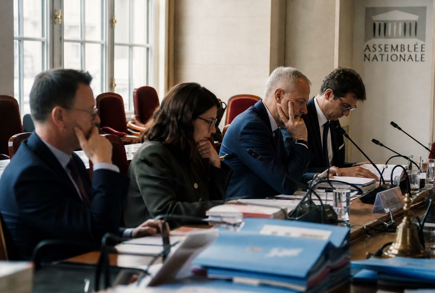 Salle de commission lumineuse avec micros de table, dossiers flous et silhouettes anonymes en séance de contrôle.