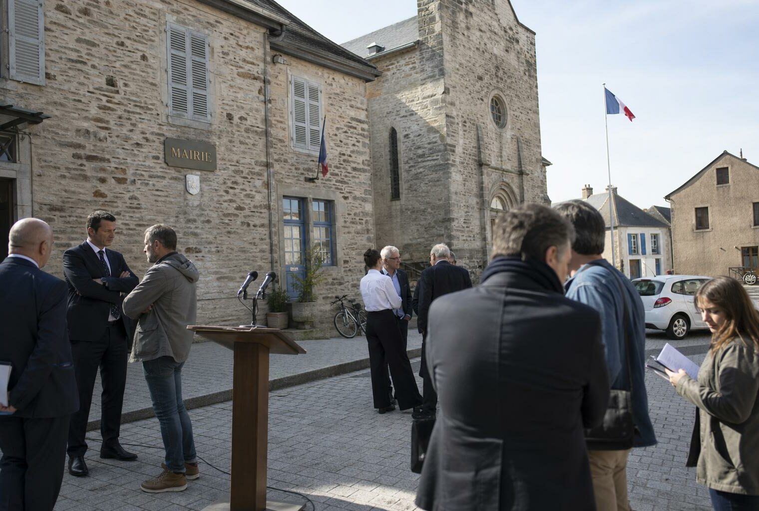 Village square in Aveyron outside a mairie, with podium microphones and a distant tricolour flag for a presidential candidacy announcement.