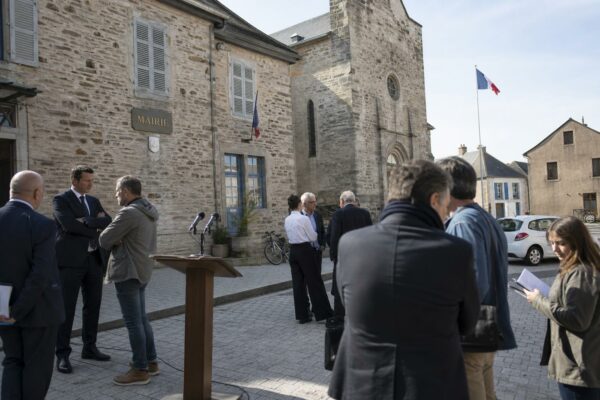 Village square in Aveyron outside a mairie, with podium microphones and a distant tricolour flag for a presidential candidacy announcement.