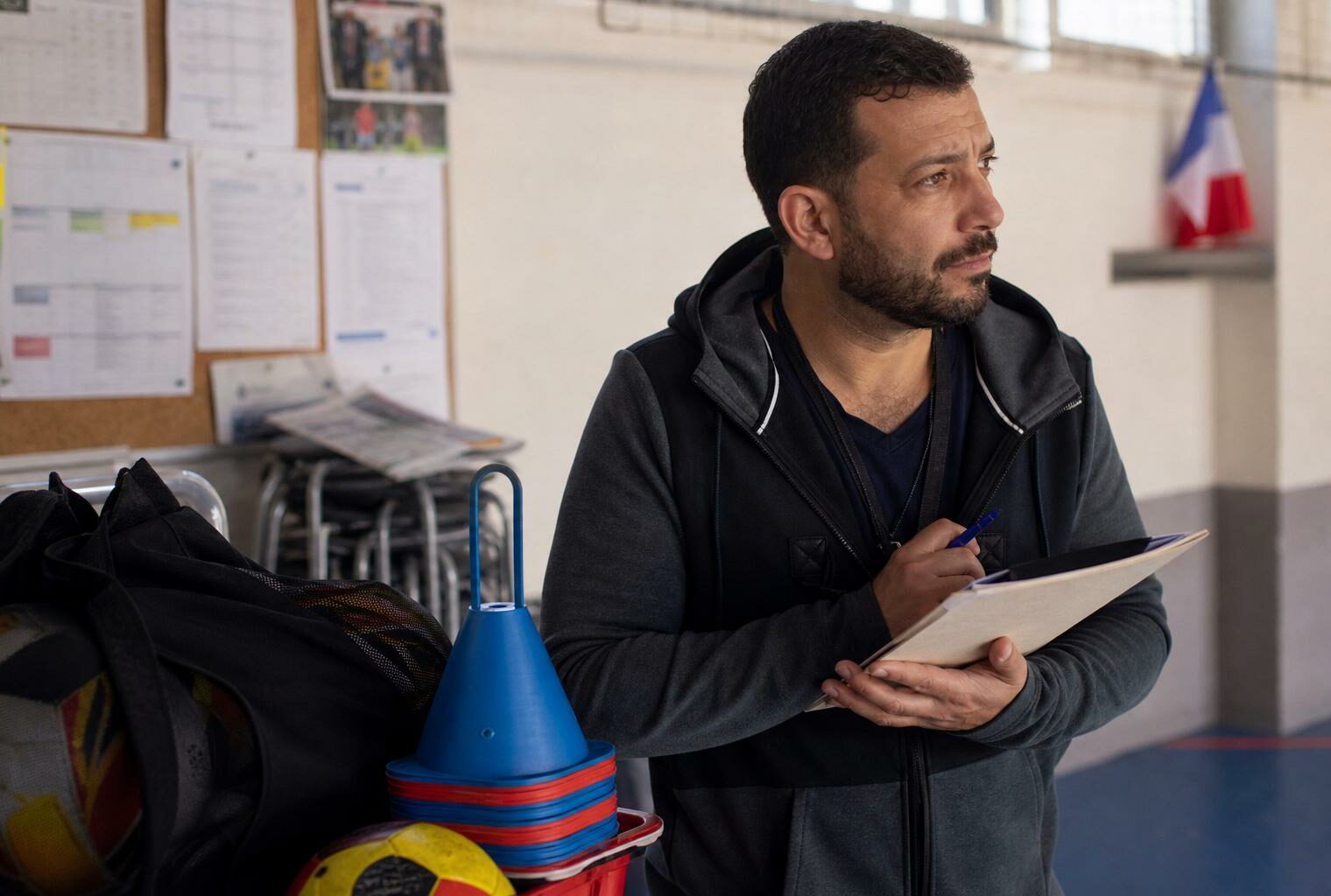 Bénévole d’un club sportif local en portrait, avec carnet et matériel de sport dans un gymnase français lumineux.