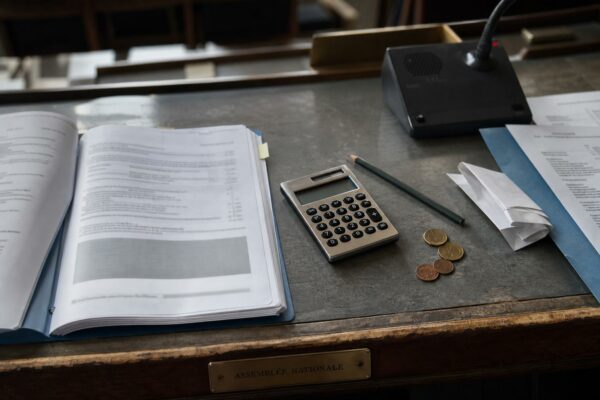 Vue en plongée d’un bureau parlementaire avec dossier budgétaire, calculatrice, pièces euro et plaque Assemblée nationale, illustrant le budget de la défense et la nomination à la Banque de France.