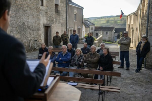 Scène de meeting politique dans un village français avec habitants assis sur des bancs et drapeau tricolore discret en arrière-plan