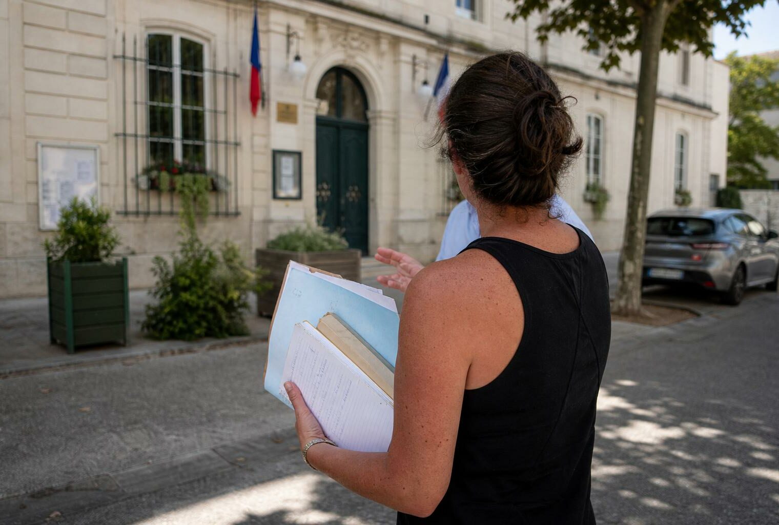 Collaborateur municipal de dos devant une mairie, carnet en main, sous la chaleur d’une journée d’été.