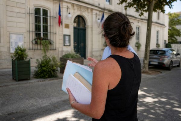 Collaborateur municipal de dos devant une mairie, carnet en main, sous la chaleur d’une journée d’été.