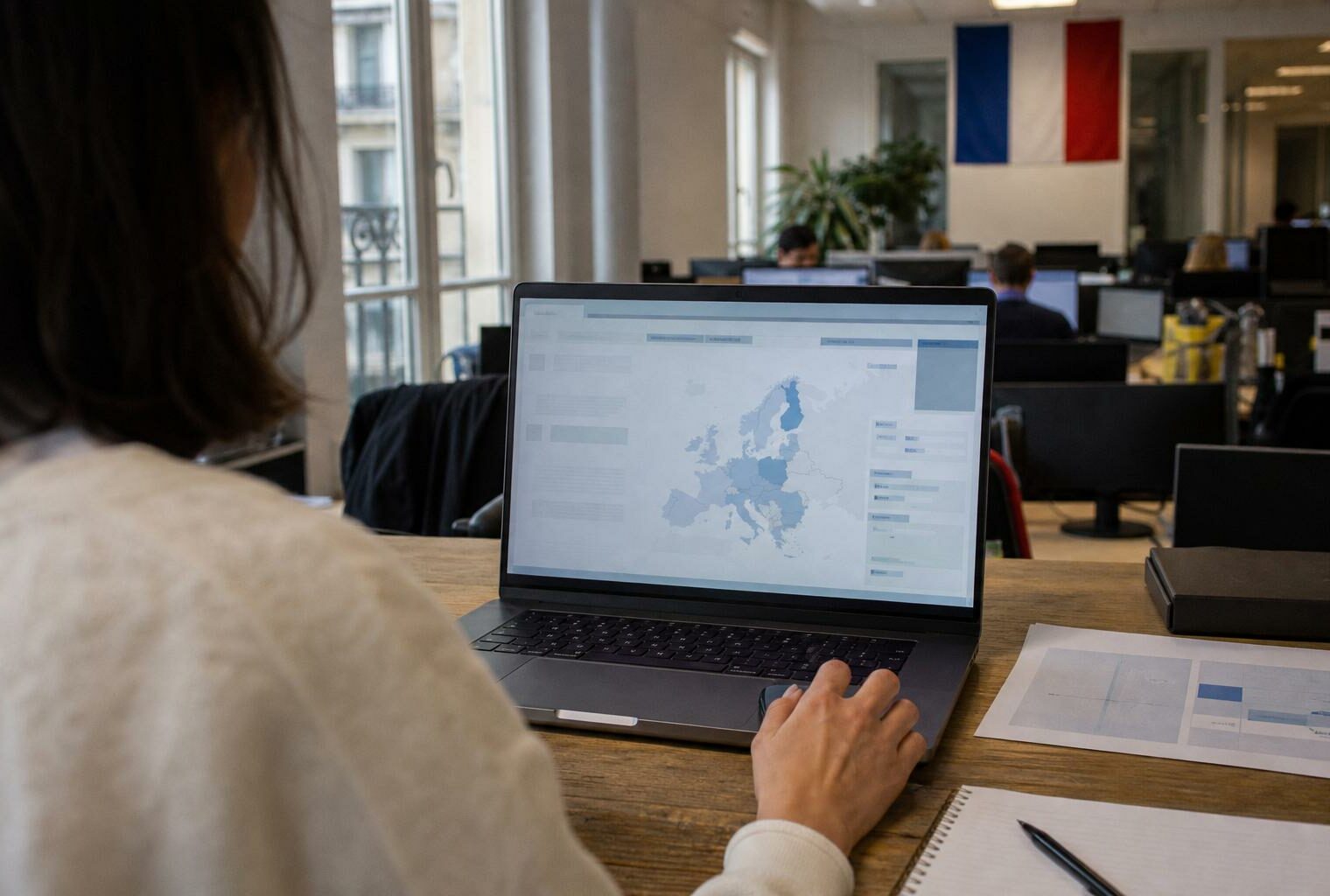 Vue par-dessus l’épaule d’un bureau de rédaction, avec ordinateur portable affichant une interface européenne abstraite.