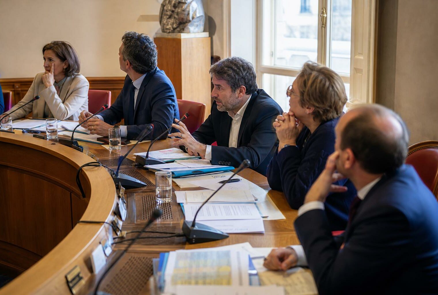 Réunion de commission parlementaire avec micros, dossiers ouverts et élus anonymes dans une salle de l’Assemblée nationale.