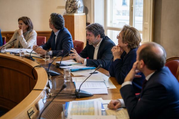 Réunion de commission parlementaire avec micros, dossiers ouverts et élus anonymes dans une salle de l’Assemblée nationale.
