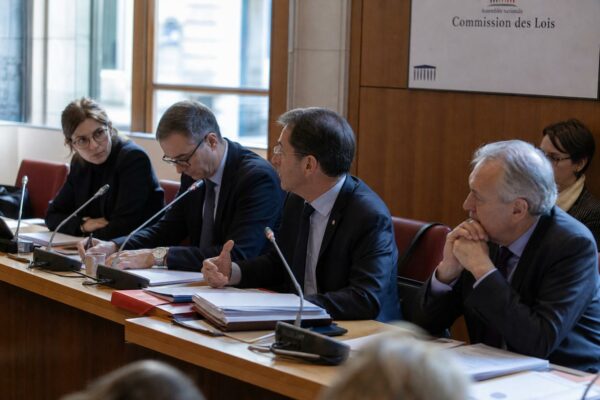 Réunion de commission parlementaire en France avec micros, dossiers ouverts et silhouettes anonymes