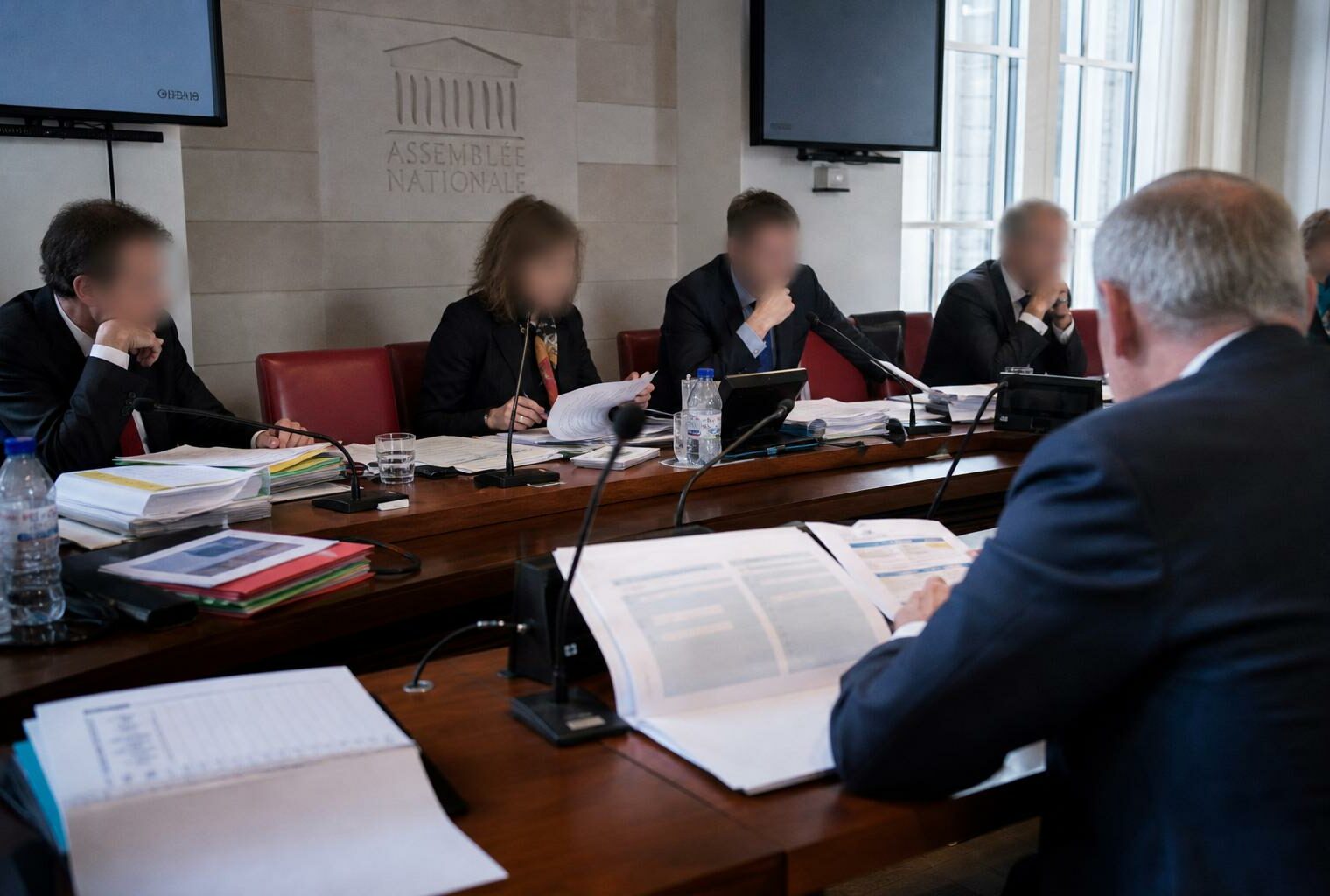 Audition parlementaire sur la Sécurité sociale avec micros, dossiers et silhouettes anonymes autour d’une table