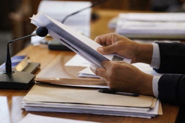 Dossier tenu à la main devant un micro de commission parlementaire, avec documents flous et lumière naturelle.