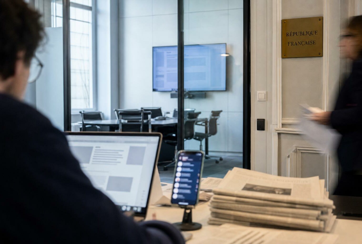 Salle de rédaction française avec écrans flous, journaux pliés et plaque institutionnelle, illustrant le débat sur la concentration des médias.