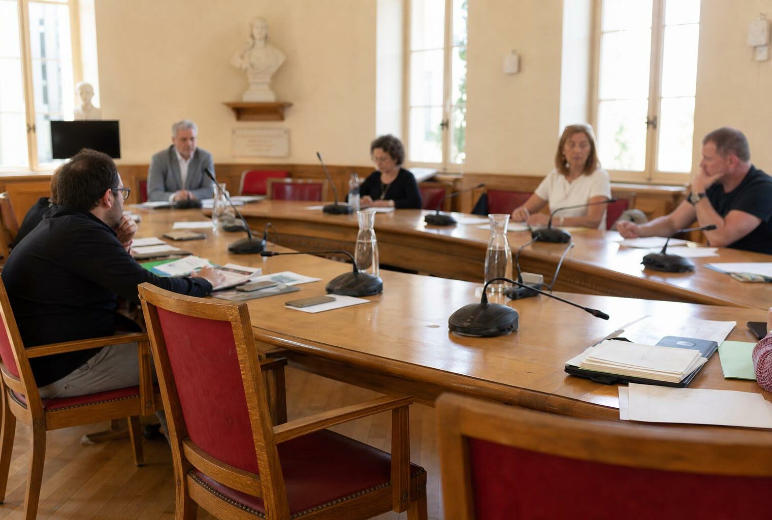 Salle municipale en plan large avec chaise vide, micros et dossiers flous lors d’un conseil local