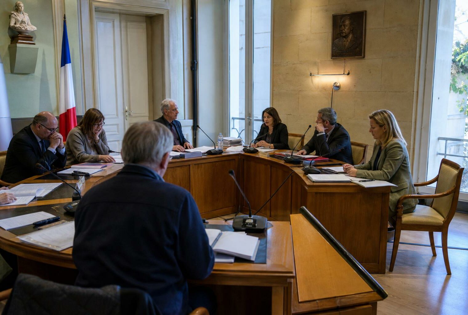Salle de conseil municipal française vue en oblique, avec micros, dossiers et une chaise vide en arrière-plan.