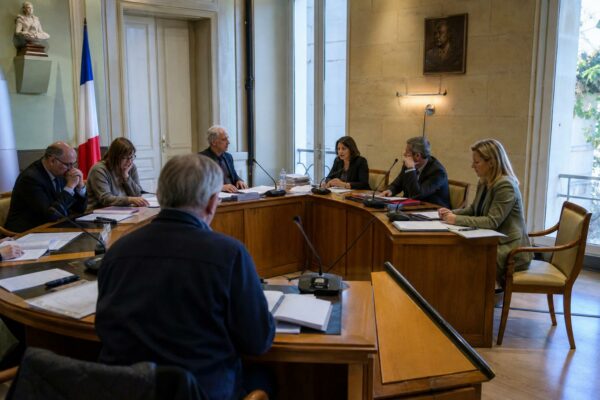 Salle de conseil municipal française vue en oblique, avec micros, dossiers et une chaise vide en arrière-plan.