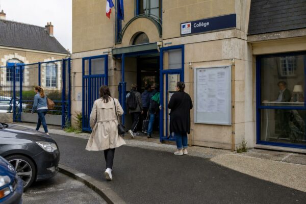 Des parents anonymes passent devant l’entrée d’un collège de ville moyenne, dans une scène de reportage claire et naturelle.