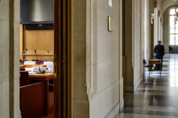 Couloir clair de l’Assemblée nationale devant une salle d’audition, ambiance institutionnelle et documentaire.