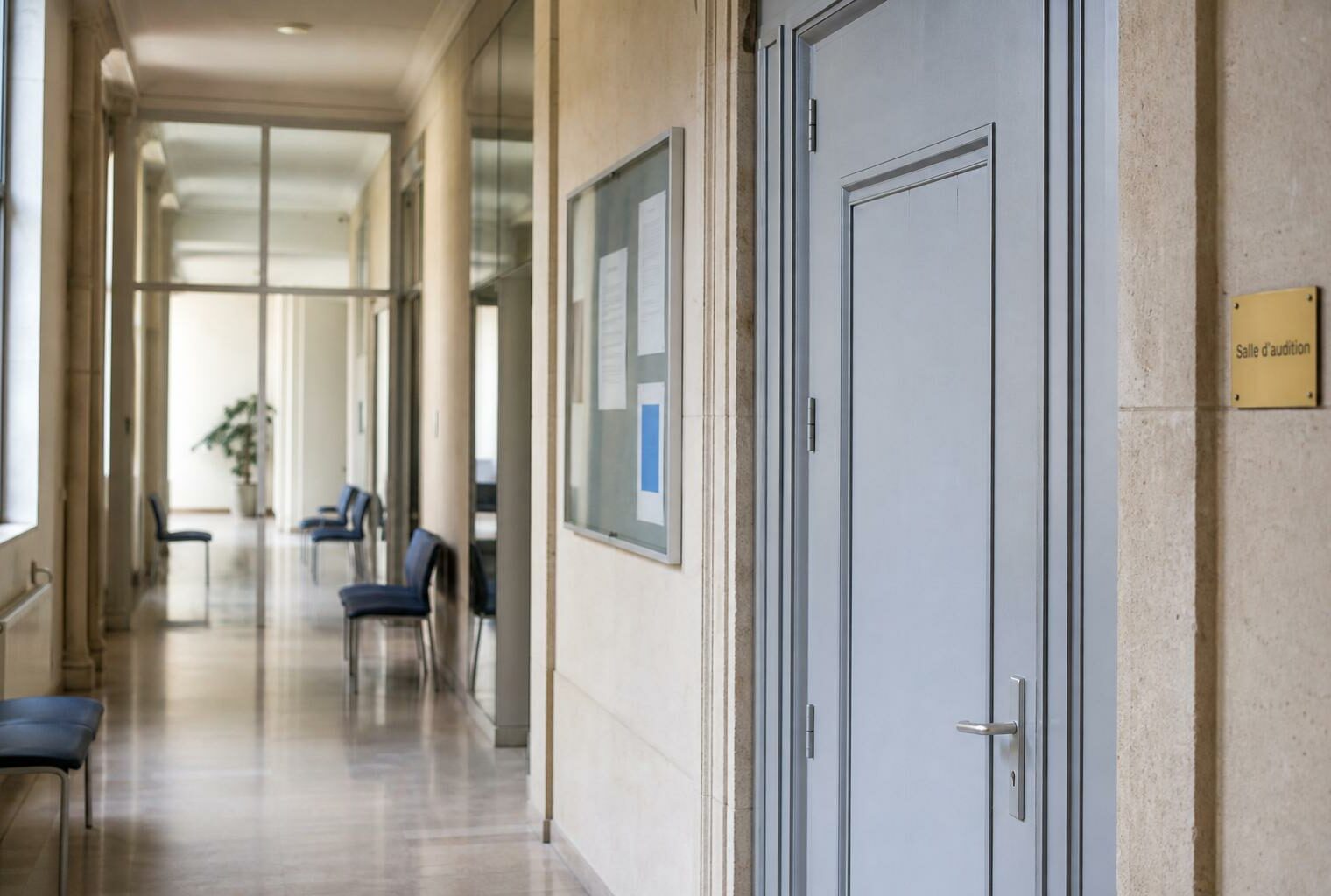 Couloir lumineux d’un bâtiment institutionnel français, avec porte sobre et salle d’audition en arrière-plan.