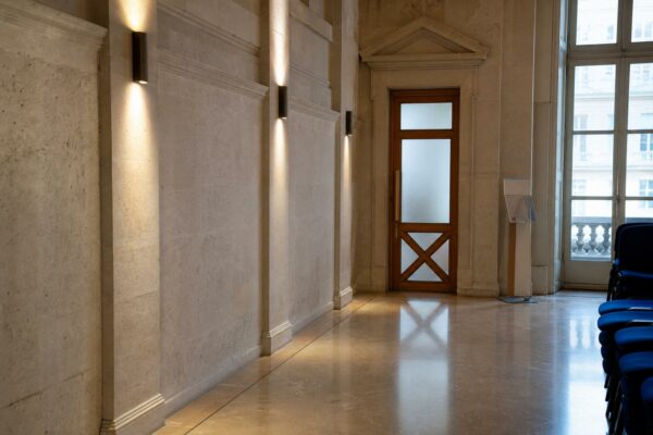 Couloir clair d’un bâtiment institutionnel français menant à une salle d’audition, ambiance sobre et documentaire.