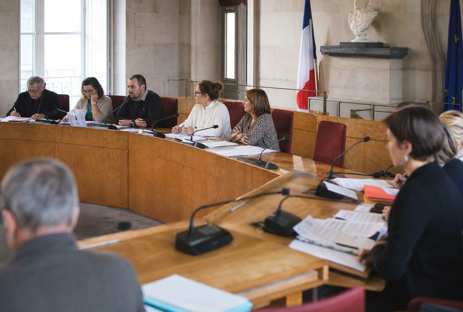 Salle municipale lumineuse avec micros, dossiers flous et une chaise vide pour illustrer le débat sur la fin de vie.