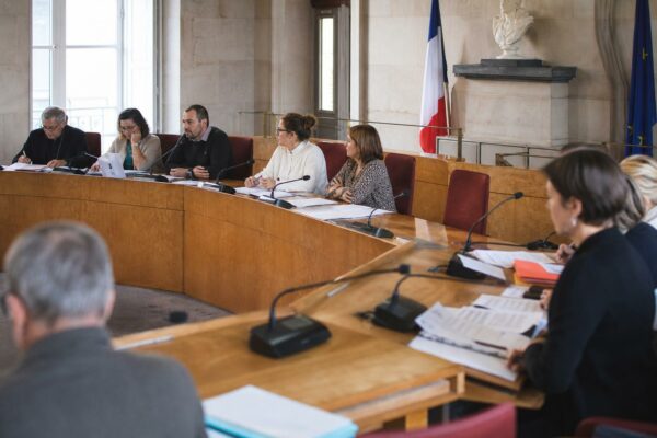 Salle municipale lumineuse avec micros, dossiers flous et une chaise vide pour illustrer le débat sur la fin de vie.