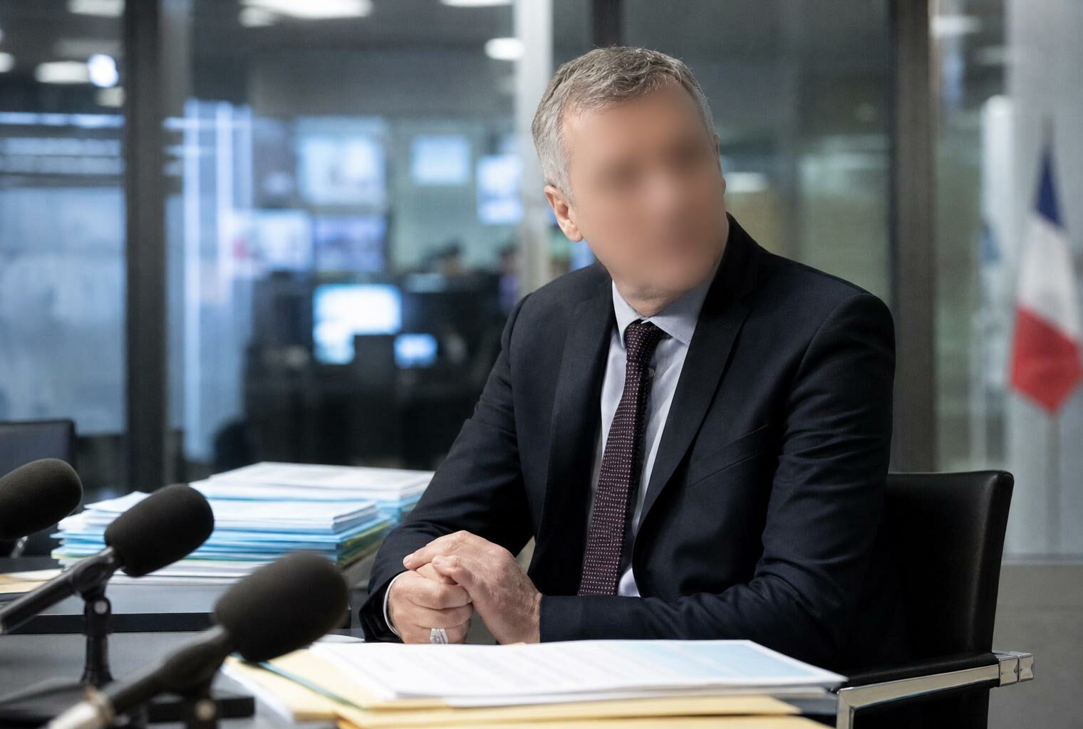Un cadre de France Télévisions dans un bureau de rédaction, avec micros de presse et drapeau tricolore flou en arrière-plan
