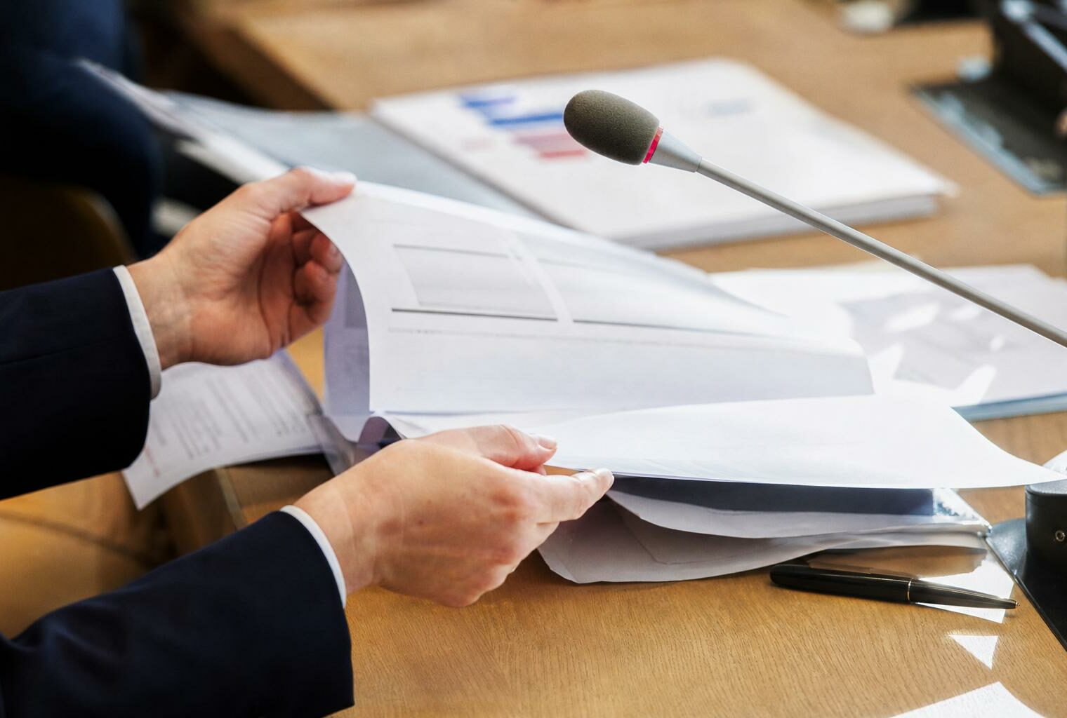 Mains anonymes autour d’un dossier et d’un micro de commission dans une salle d’audition parlementaire lumineuse