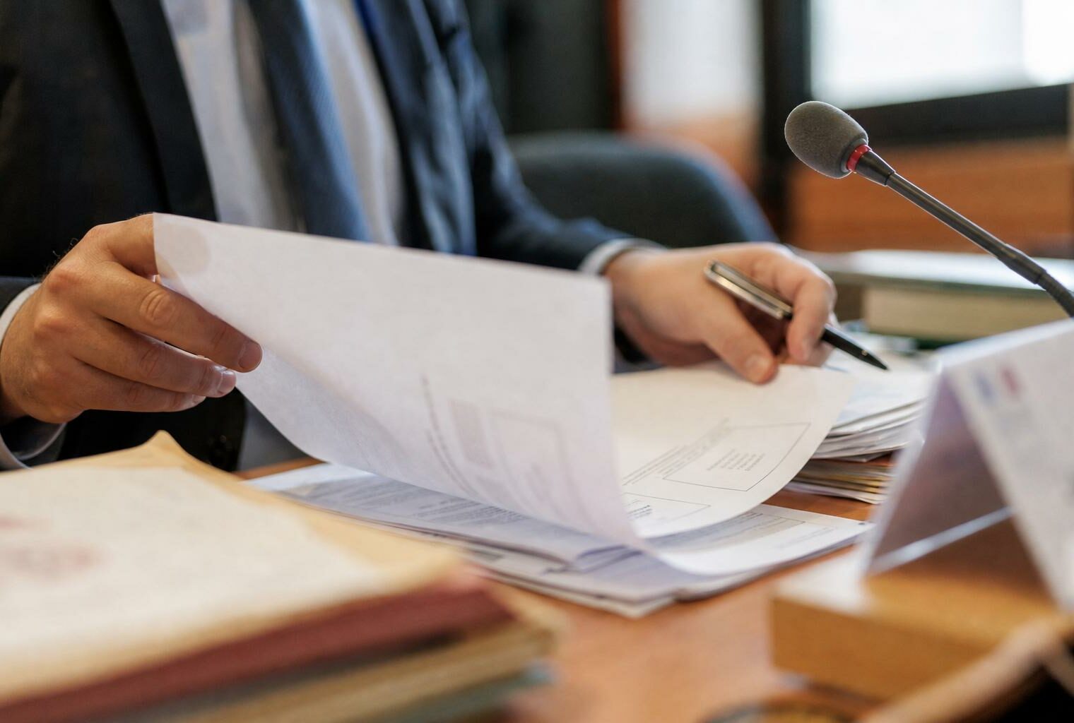Dossier administratif flou et micro de commission sur une table, main anonyme en train de feuilleter un document.