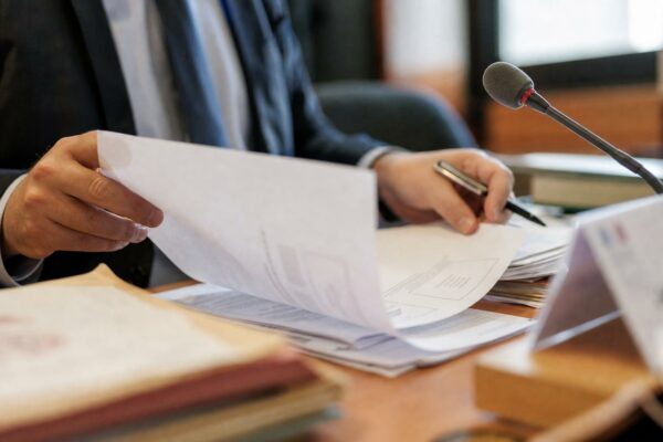 Dossier administratif flou et micro de commission sur une table, main anonyme en train de feuilleter un document.
