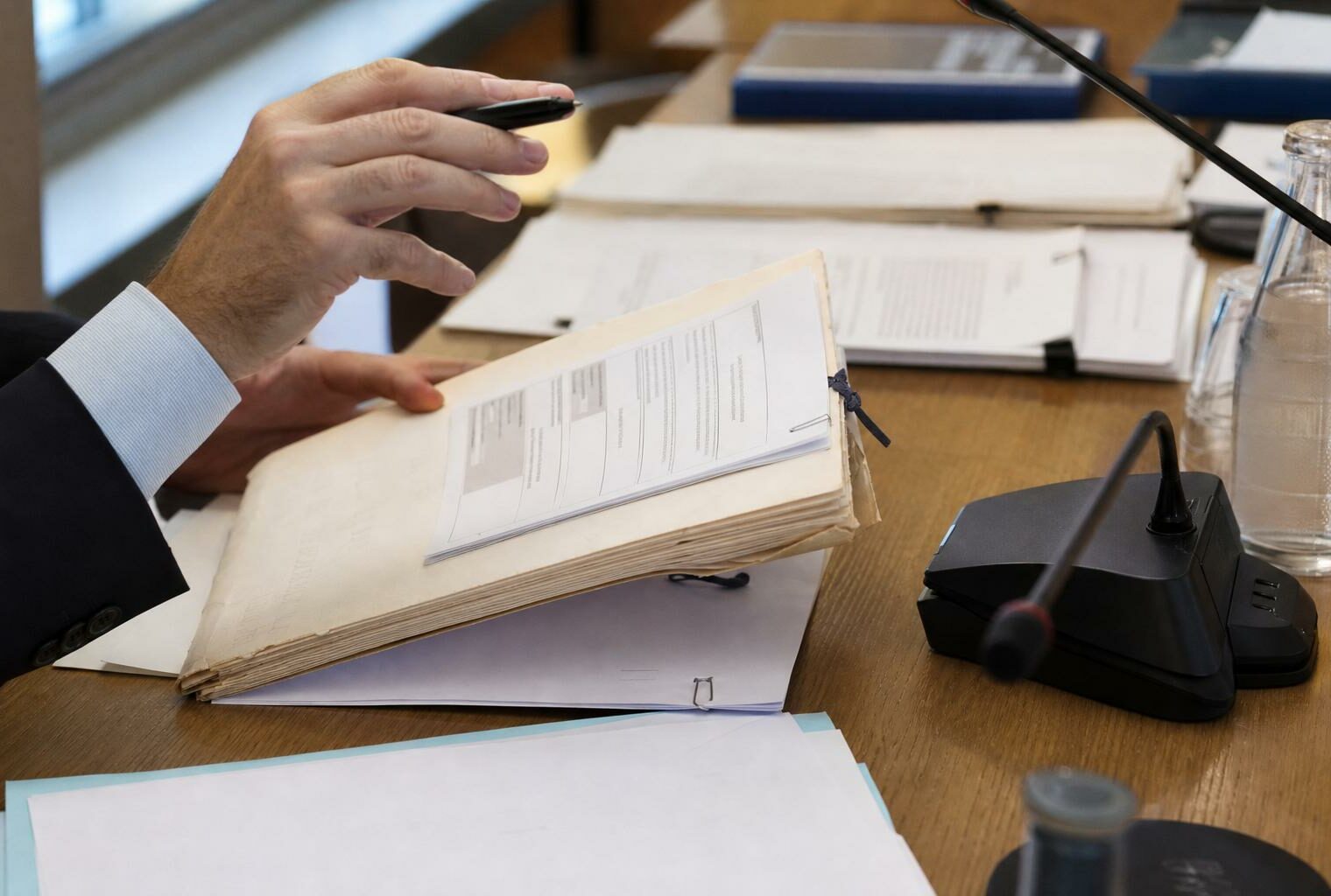 Mains anonymes consultant un dossier de nomination près d’un micro de commission dans une salle institutionnelle lumineuse.