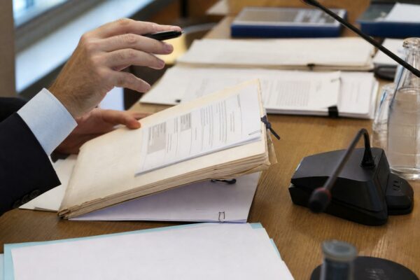 Mains anonymes consultant un dossier de nomination près d’un micro de commission dans une salle institutionnelle lumineuse.