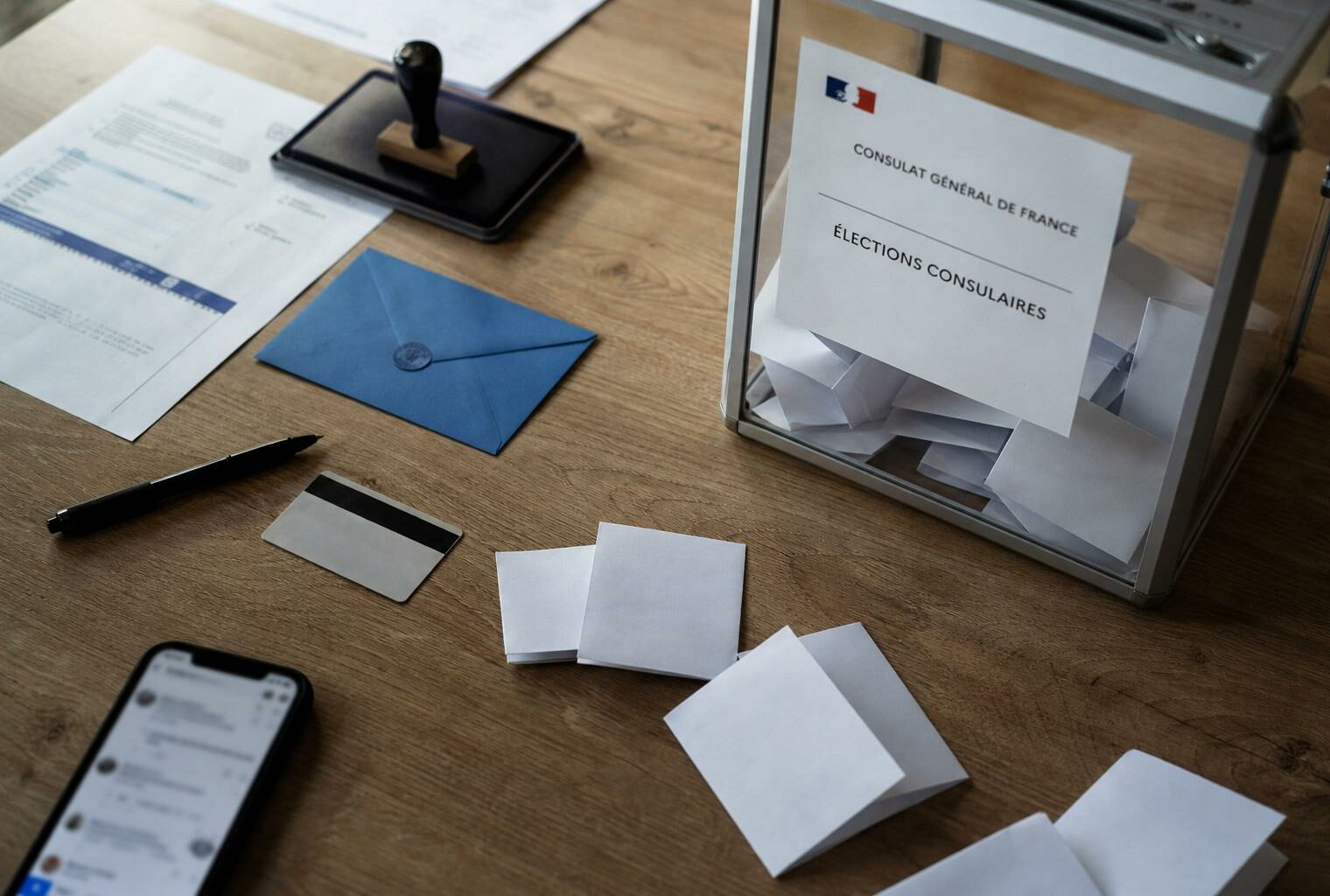 Vue en plongée d’un bureau de vote consulaire avec urne transparente, bulletins pliés et documents administratifs flous.