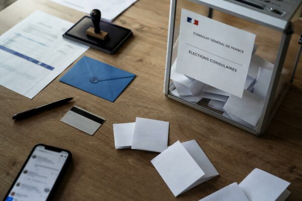 Vue en plongée d’un bureau de vote consulaire avec urne transparente, bulletins pliés et documents administratifs flous.