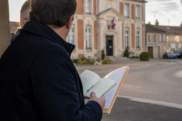 Un élu local de dos consulte un carnet devant une mairie de village, dans une lumière douce de fin d’après-midi.