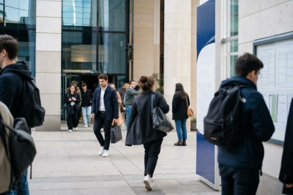 Des étudiants anonymes traversent l’entrée lumineuse d’un campus parisien de grande école, en scène de vie quotidienne.