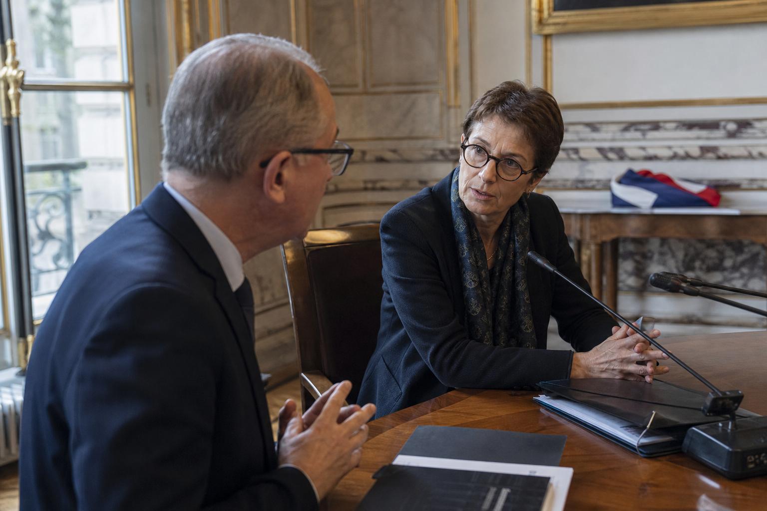 Deux responsables en réunion bilatérale dans un salon ministériel parisien, avec marbre, dorures et dossiers flous.