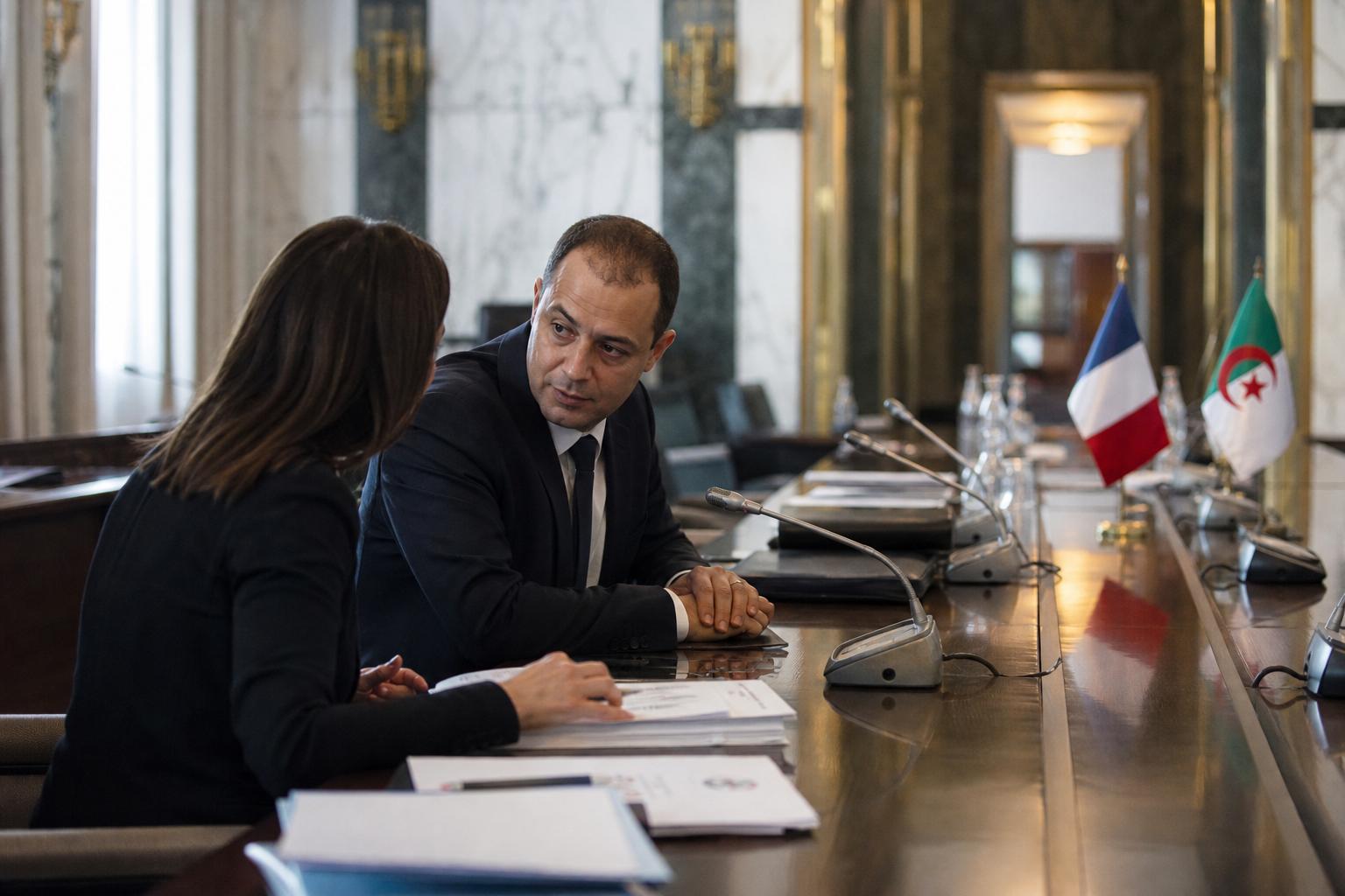 Deux responsables anonymes discutent dans une salle institutionnelle avec micros et petits drapeaux français et algérien.