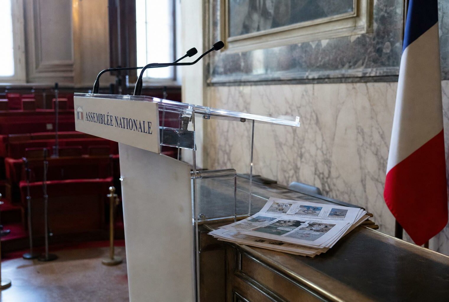 Salle de presse de l’Assemblée nationale avec pupitre vide, micros et pages de bande dessinée, en contexte journalistique.