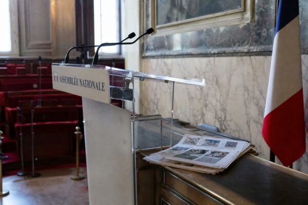 Salle de presse de l’Assemblée nationale avec pupitre vide, micros et pages de bande dessinée, en contexte journalistique.