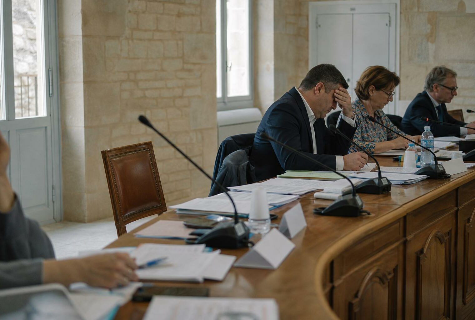 Salle du conseil municipal avec chaise vide, micros et dossiers flous dans une ambiance claire de reportage