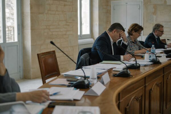 Salle du conseil municipal avec chaise vide, micros et dossiers flous dans une ambiance claire de reportage