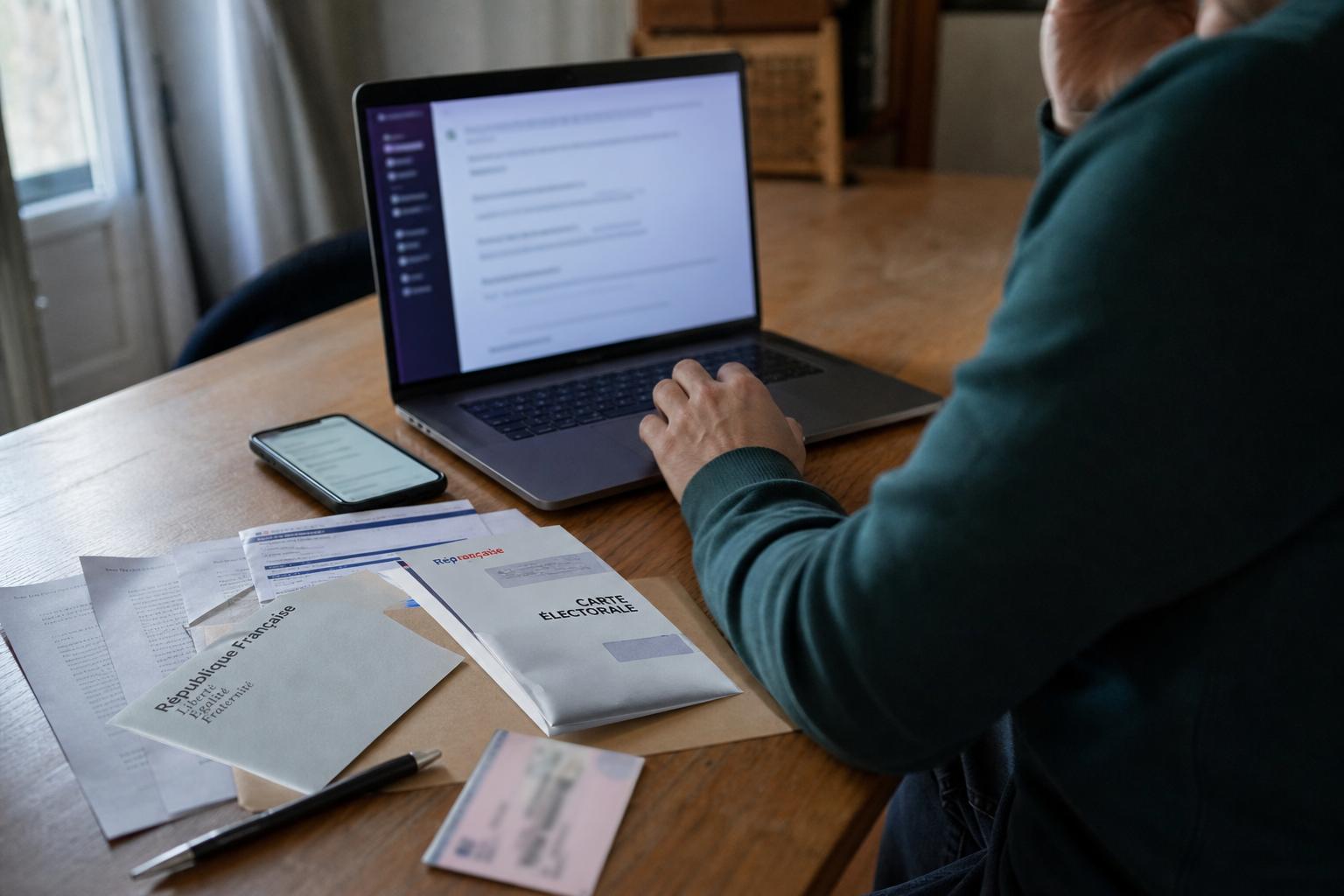 Un électeur consulte une interface IA sur ordinateur et smartphone avec des documents de vote flous sur une table.