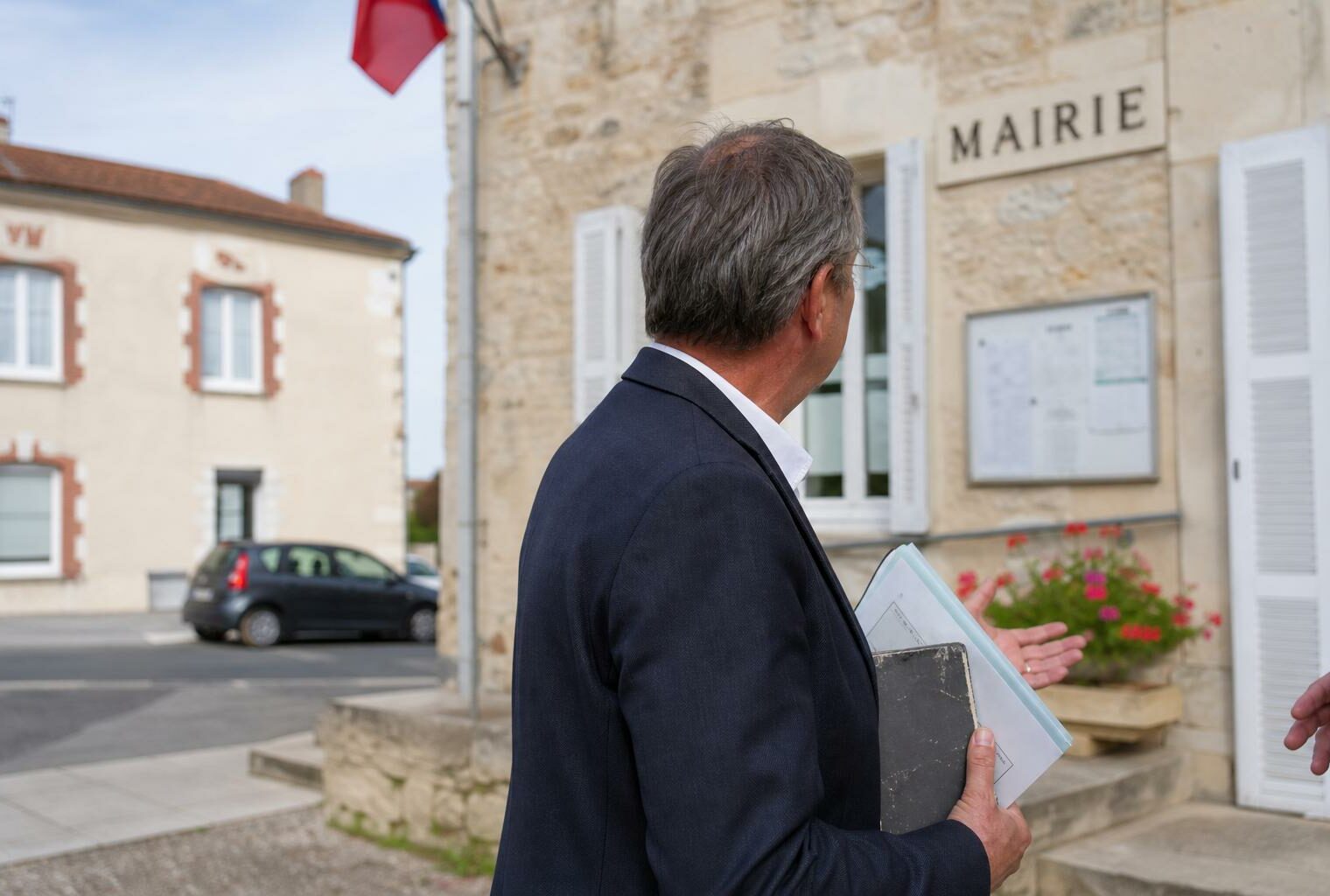 Un élu local anonyme tient un dossier devant une mairie de village, dans une scène de portrait journalistique lumineuse.