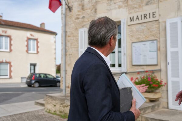 Un élu local anonyme tient un dossier devant une mairie de village, dans une scène de portrait journalistique lumineuse.
